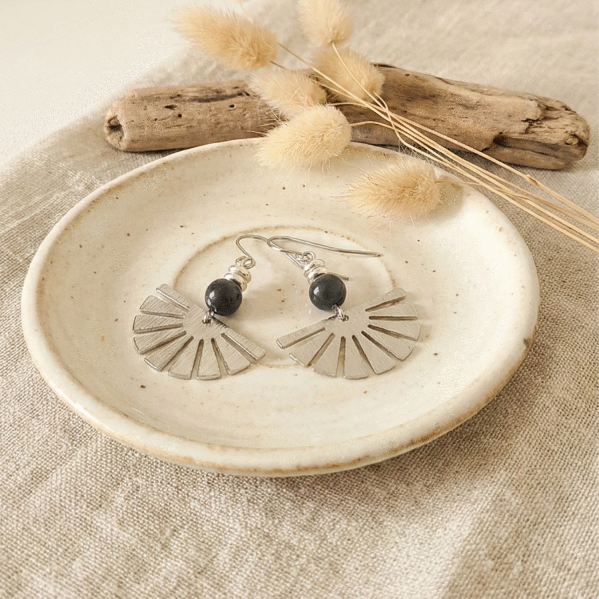 photo de boucles d'oreilles pendantes forme éventail argenté avec perle noire sur rondin de bois